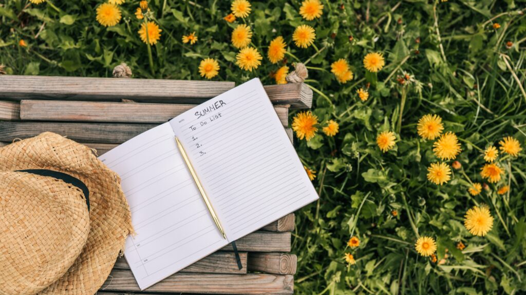 Open notebook with "Summer To Do List" written inside, resting on a wooden bench in a field of blooming dandelions, with a straw hat placed nearby in soft afternoon sunlight.
